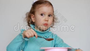 一个漂亮的小女孩坐在桌子旁，用盘子里的勺子吃食物。 这孩子仔细咀嚼。 早餐
